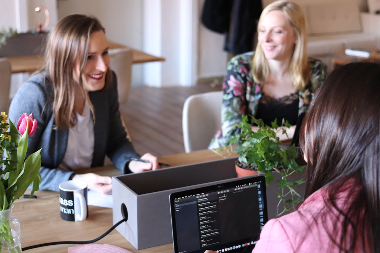 our-services-3 Three women engaging in a casual business meeting in a modern office setting with laptops and plants.